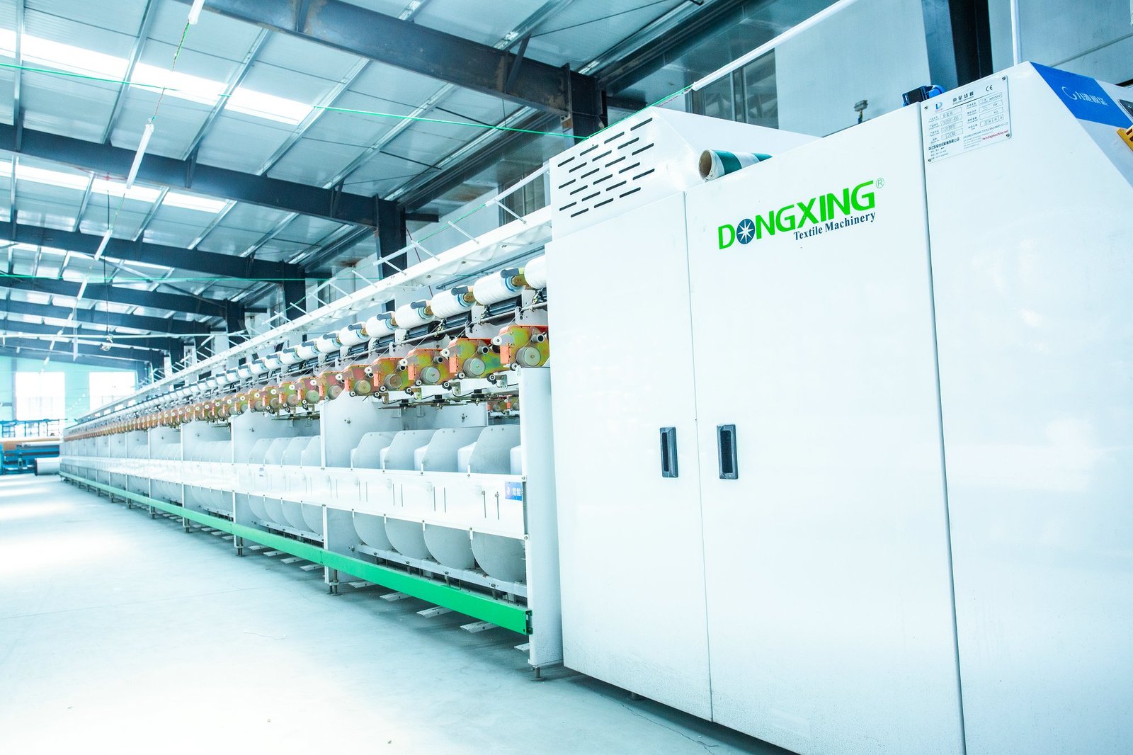 A bright, spacious textile factory interior featuring a large, advanced weaving machine designed for manufacturing concrete mattresses. The machine is lined with spools of synthetic fibers, essential for creating durable products. The clean, industrial environment showcases the efficiency of concrete mattress production, with ample natural light illuminating the space.