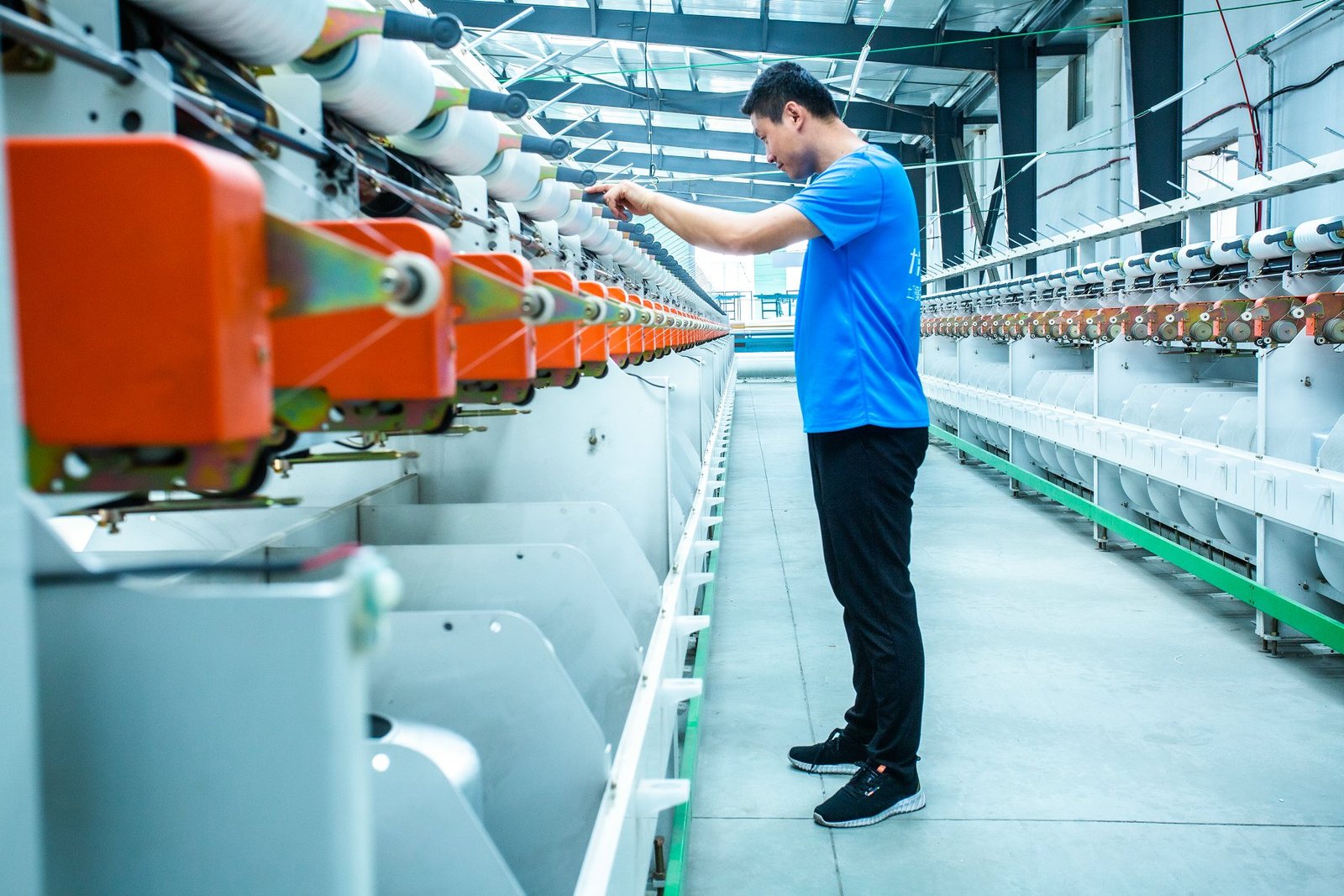 A worker in a blue shirt operates a large weaving machine in a well-lit factory. The machine features multiple spools of thread, showcasing the intricate process of fabric creation used for concrete mattresses. The clean and organized workspace highlights the industrial environment of production for durable concrete products.