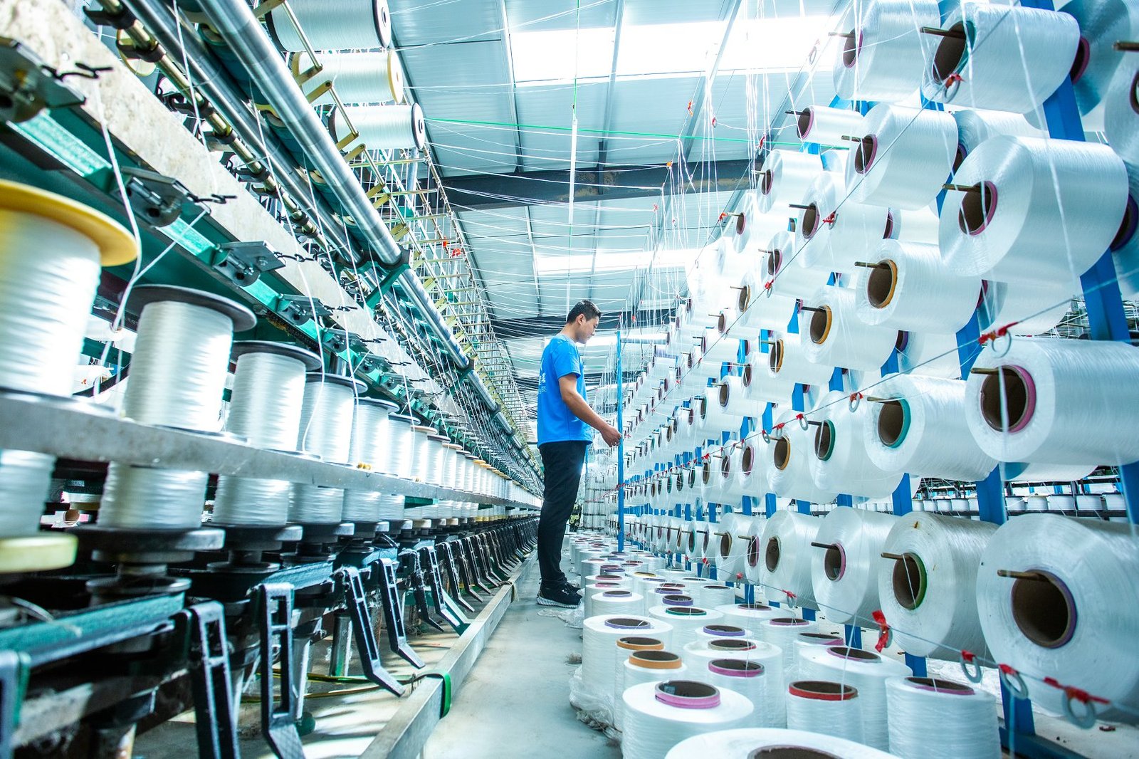 A production worker in a blue shirt is surrounded by rows of spools of synthetic fibers, essential for creating articulated concrete mattresses. The bright and organized factory interior showcases the weaving machinery, highlighting the intricate process of textile manufacturing. This image reflects the commitment to quality materials in producing durable concrete mattresses.