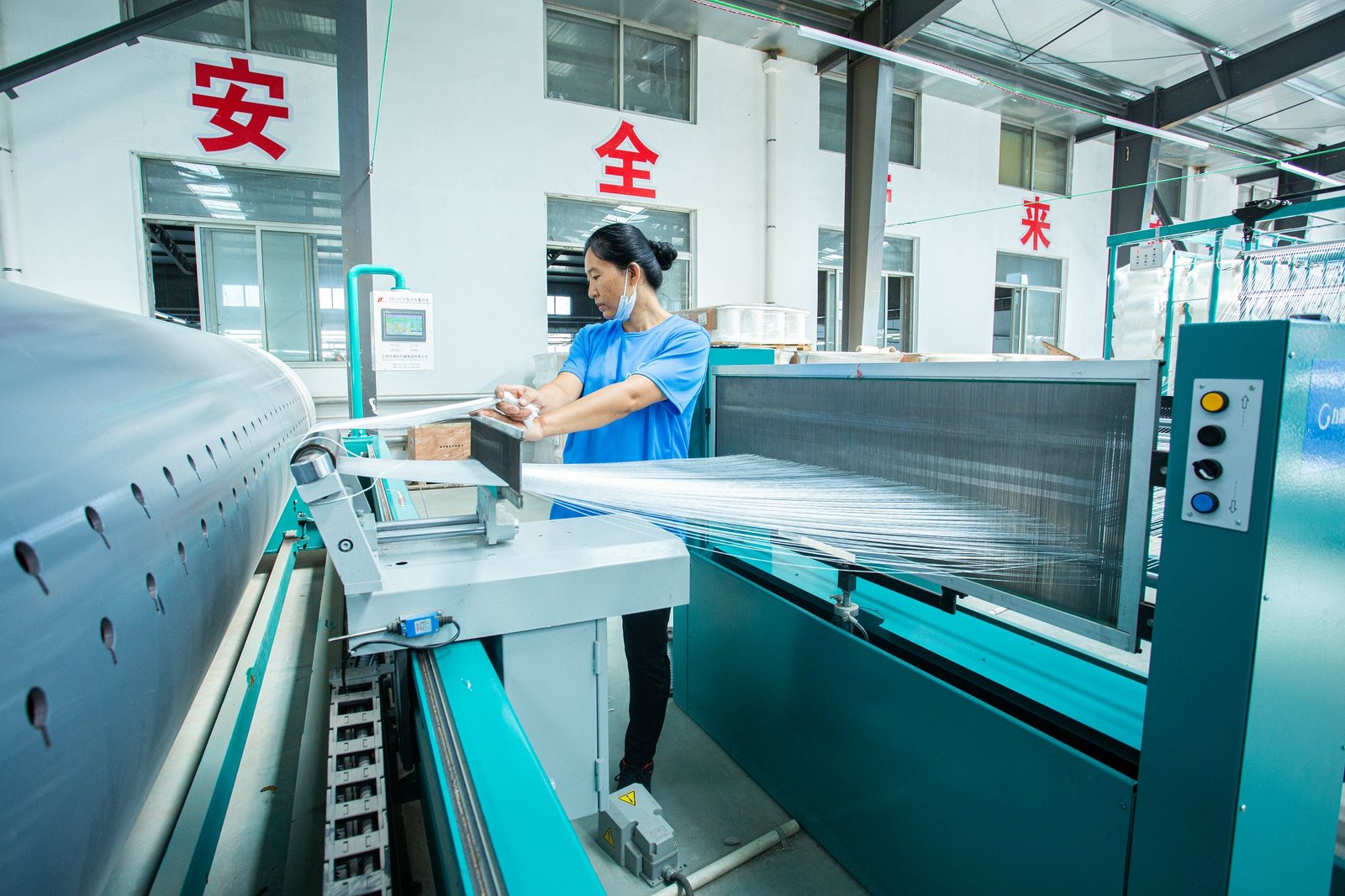 A factory worker operates machinery in a bright manufacturing facility, weaving synthetic fibers used for producing articulated concrete mattresses. The machine is processing strands of material, showcasing the industrial environment with clear windows and organized workspaces.