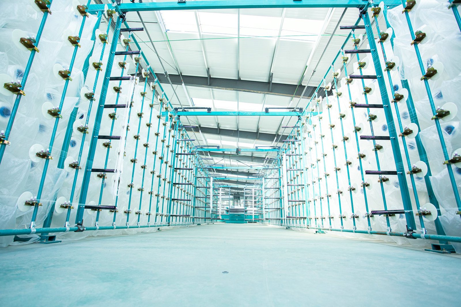 View of an industrial workshop featuring large weaving machines and framework for manufacturing articulated concrete mattresses. The space is bright with natural light filtering through the ceiling, showcasing multiple turquoise metal frames and white fabric rolls on either side, indicating an advanced manufacturing process for durable concrete products.