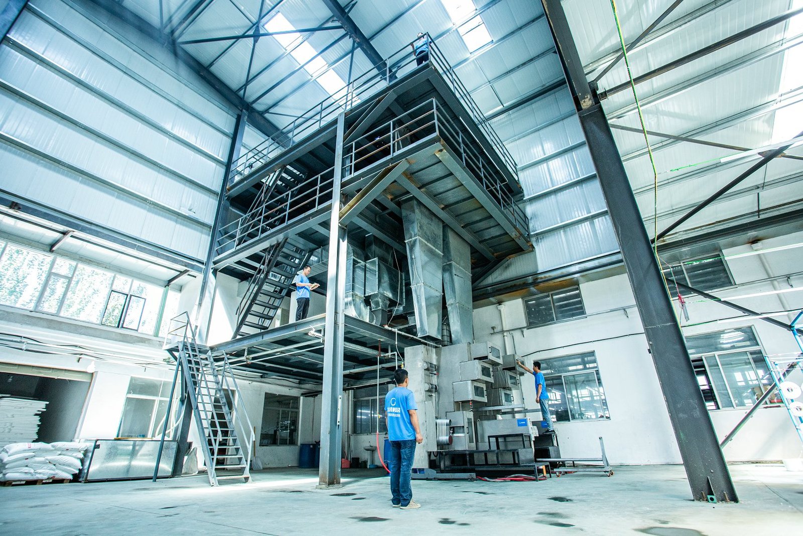 A spacious concrete mattress manufacturing facility showcasing modern machinery. Workers are actively engaged on different levels of a multi-tiered platform, with bright industrial lighting illuminating the environment. The setting includes large, stainless steel equipment used in the production of concrete mattresses, as well as a clean floor space indicative of a well-organized factory. This image represents the cutting-edge technology involved in producing durable and effective concrete mattresses.