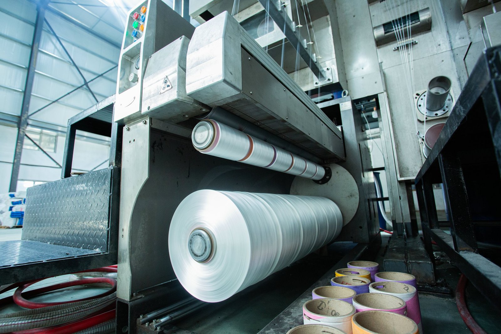 Close-up view of industrial machinery in a concrete mattress manufacturing facility. The image captures a large spool of white, synthetic fabric being processed, showing the rhythmic operation of the machine. Adjacent are colorful paper rolls, used to collect materials or assist in the production process, set against a bright, industrial backdrop.