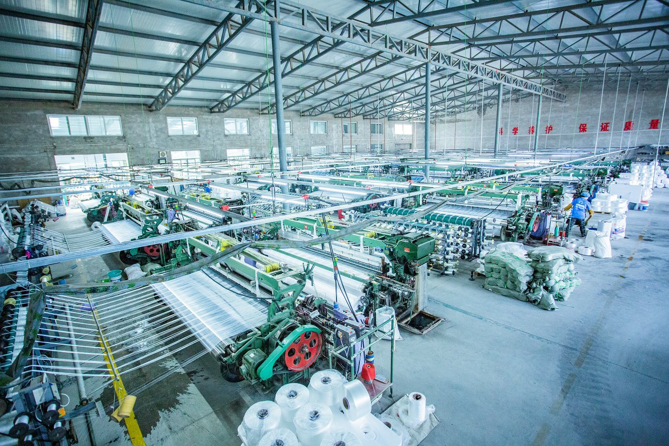 An expansive view of the interior of a concrete mattress production factory, showcasing an array of weaving machines in action. The machinery features vivid green and red components while rolls of synthetic fibers and woven fabrics lie ready for processing. High ceilings and large windows illuminate the industrial setting, where workers can be seen engaged in the manufacturing process, demonstrating the scale of operation involved in producing durable concrete mattresses.