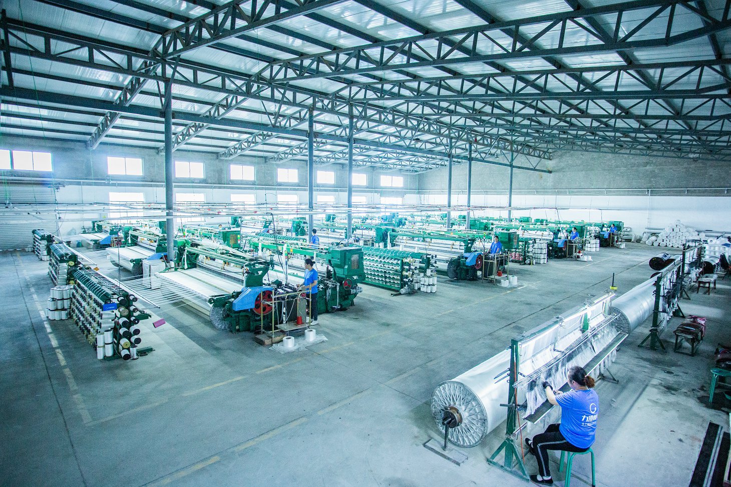 A bustling factory interior showcasing the production of concrete mattresses. Large weaving machines dominate the space, with employees in blue shirts operating them. The factory's high ceilings and organized layout are visible, alongside rolls of various fabrics stacked orderly. Sunlight streams through the windows, illuminating the industrial environment, emphasizing the extensive scale and efficiency of concrete mattress production.